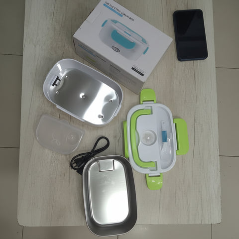 An electric heating lunch box set laid out on a light wood floor, including a stainless steel heating pan, a white and green insulated container, a clear lid, a power cord, and the product box.
